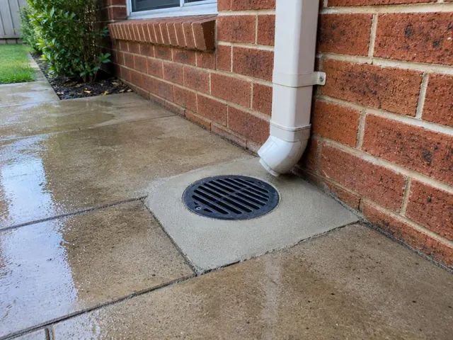 Outdoor gully grate beside a brick wall and downpipe on wet paving in Ropes Crossing